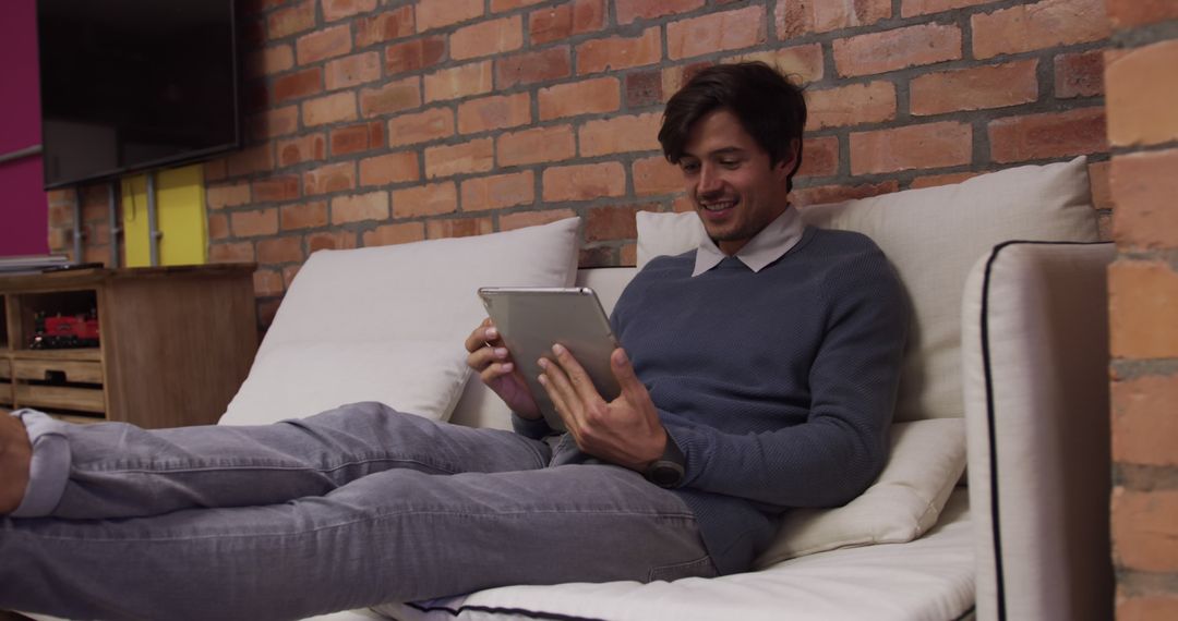 Young Man Relaxing on Sofa with Tablet in Modern Brick Wall Room - Free Images, Stock Photos and Pictures on Pikwizard.com