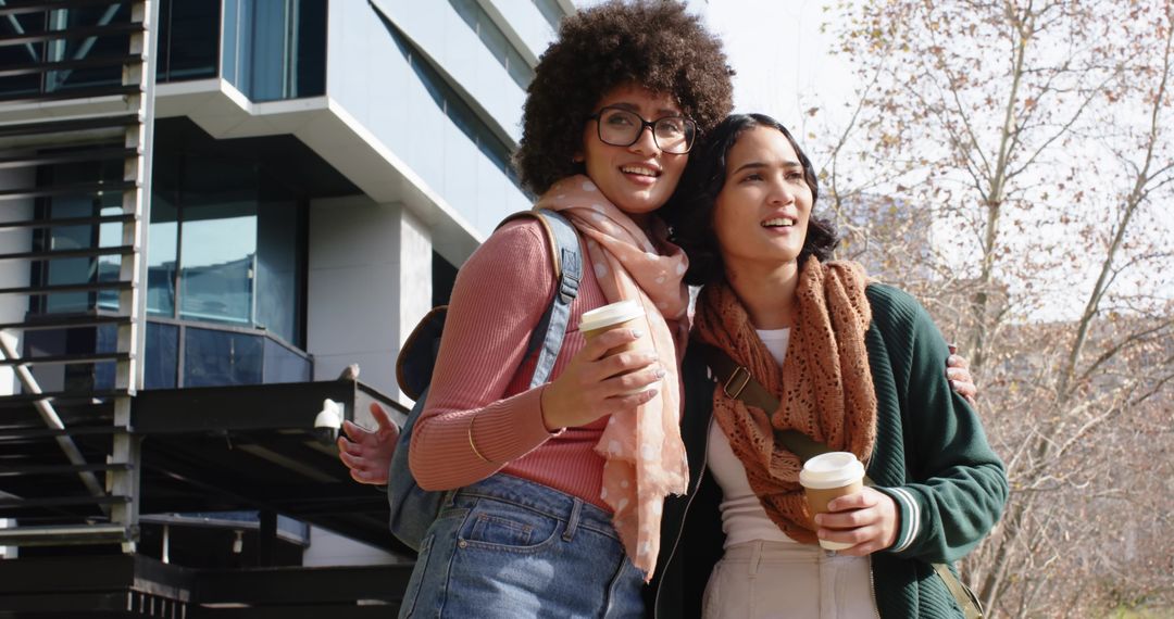 Young women enjoying coffee and laughing in urban campus with backpacks and scarves
