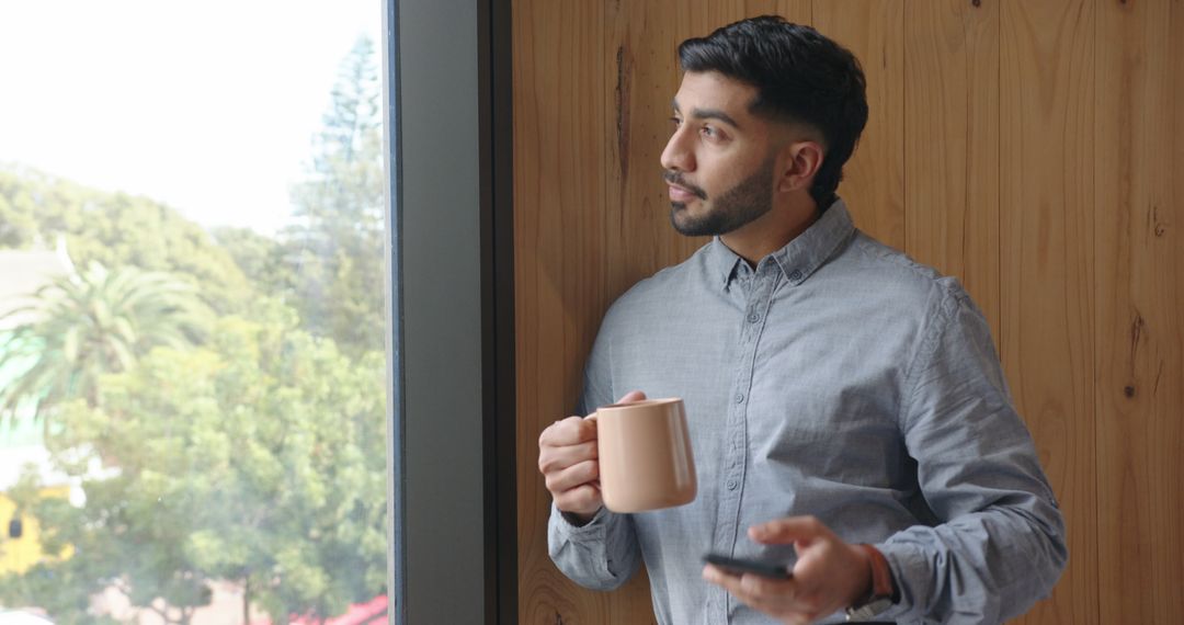 Young Man Reflecting by Window with Coffee and Phone Indoors