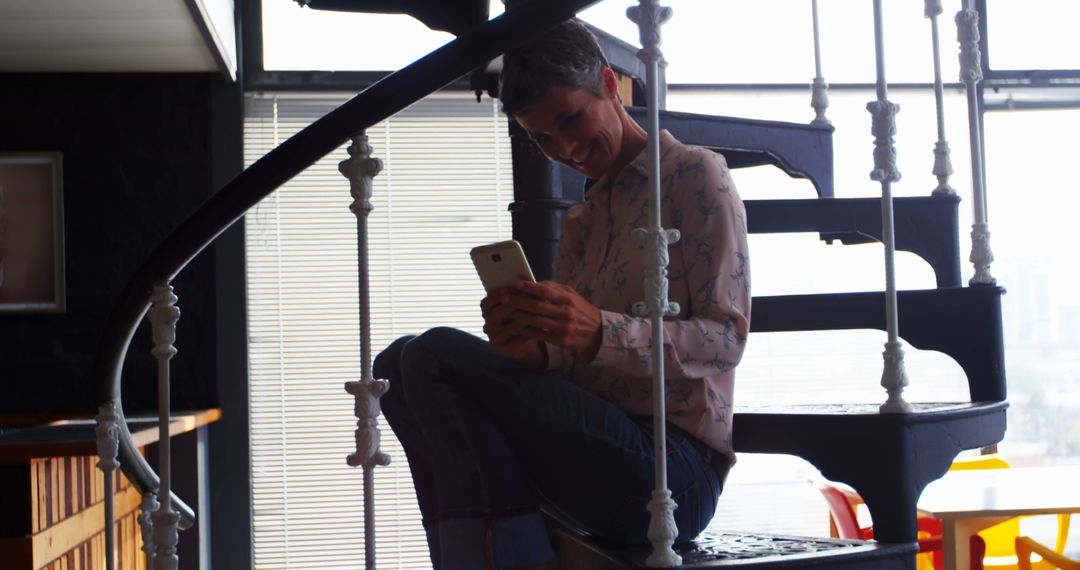 Man sitting on spiral staircase smiling while using smartphone - Free Images, Stock Photos and Pictures on Pikwizard.com