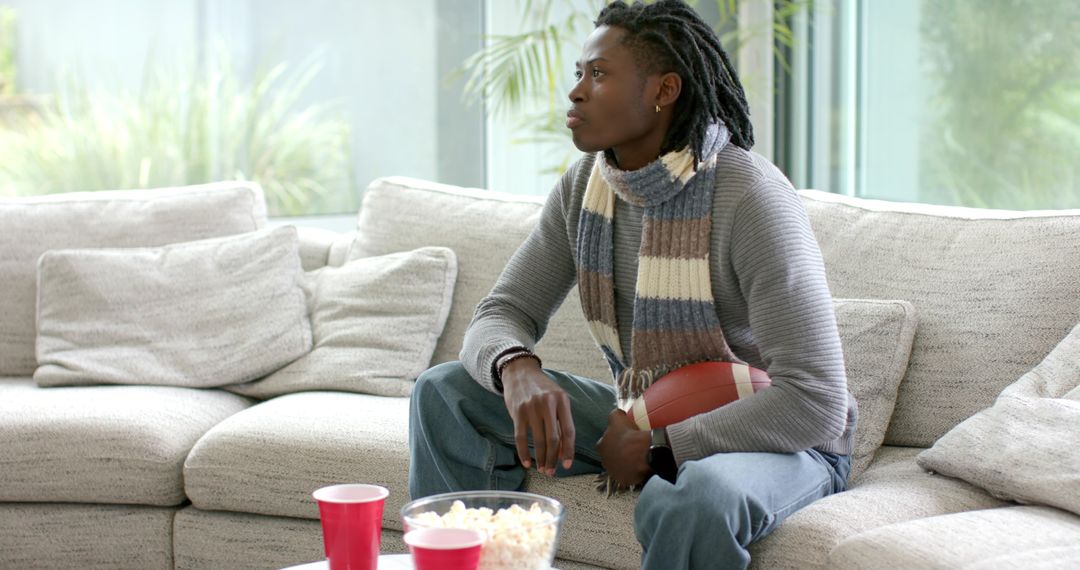 Casual man holding football watching football game on couch with popcorn, red cups and scarf
