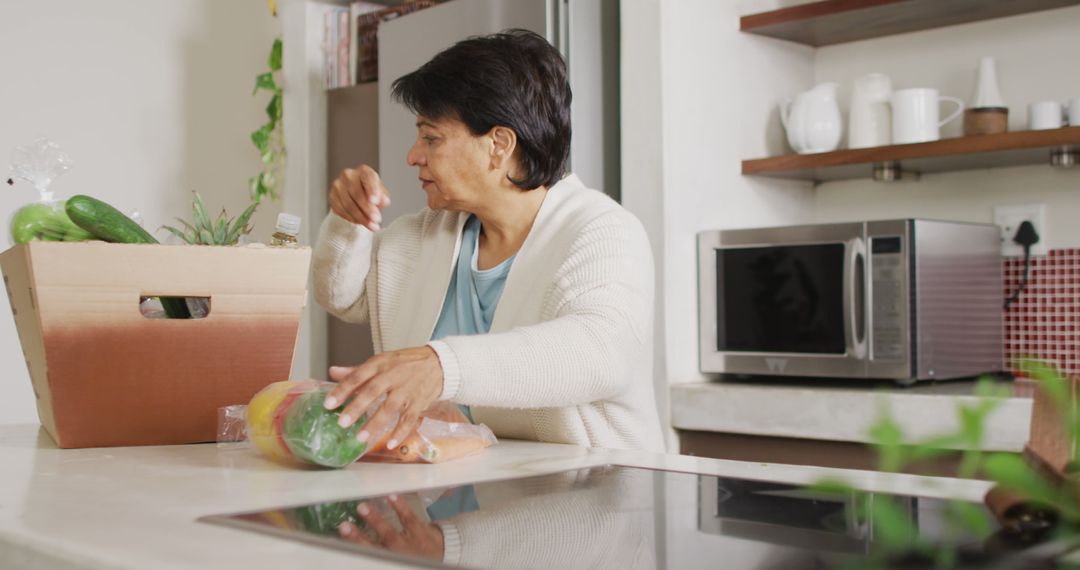 Senior biracial woman grabbing vegetables from box in kitchen alone - Free Images, Stock Photos and Pictures on Pikwizard.com