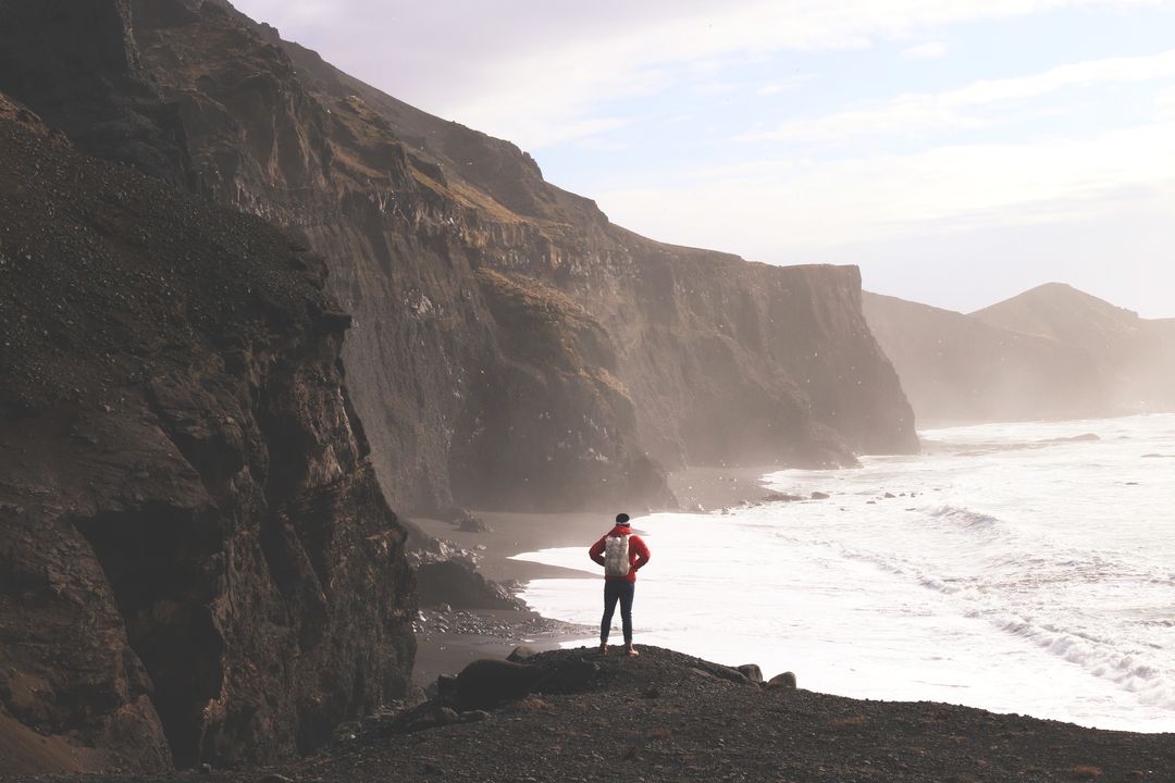 Solo hiker standing on black-sand cliff overlooking dramatic volcanic coastline and crashing waves