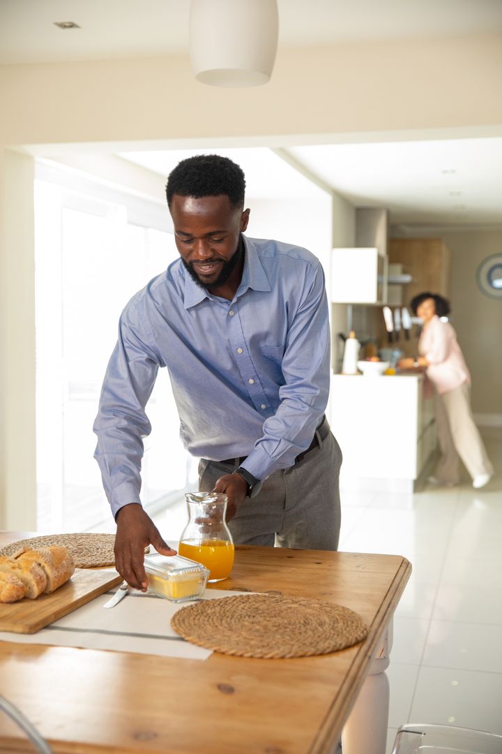 Man Preparing Breakfast Table at Home with Partner in Background