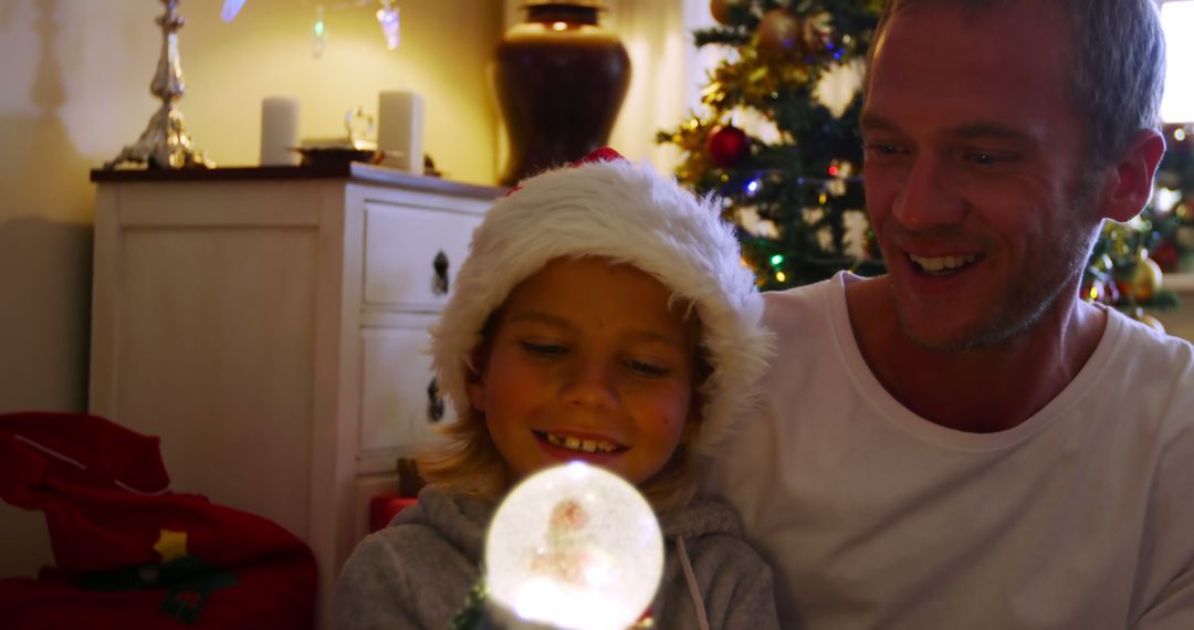 Father and Son Enjoying Christmas Snow Globe by Tree - Free Images, Stock Photos and Pictures on Pikwizard.com