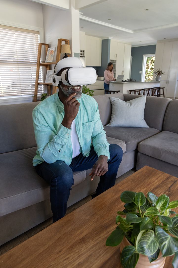 Man Wearing VR Headset on Sofa While Partner Working on Laptop in Modern Open-Plan Living Room