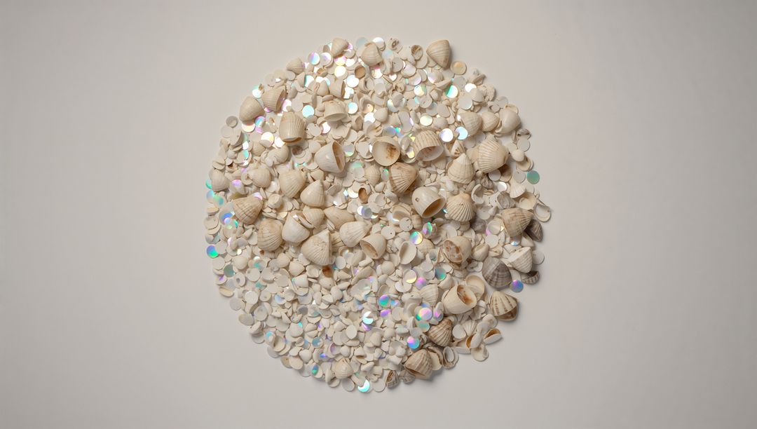 Forming circular pile of pale seashells and holographic sequins on cream tabletop