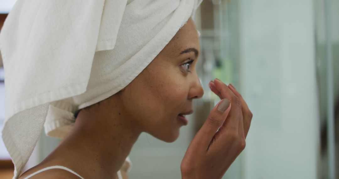 Biracial woman wearing towel on head applying cream on her face - Free Images, Stock Photos and Pictures on Pikwizard.com