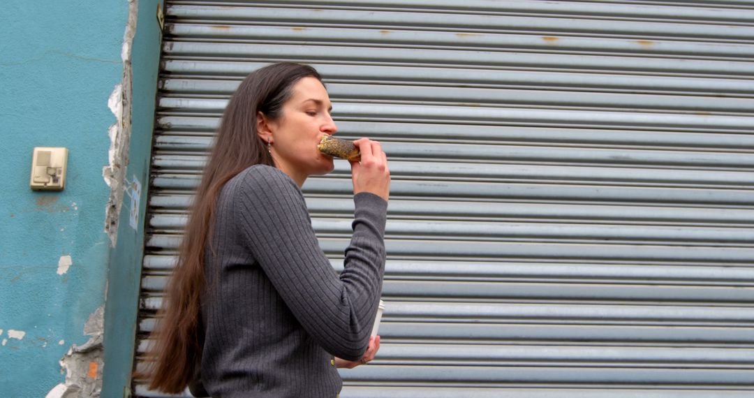 Woman with long brown hair enjoying ice cream by metal shutter - Free Images, Stock Photos and Pictures on Pikwizard.com