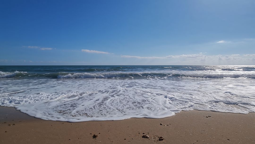 Sunlit ocean waves rolling onto sandy beach with foamy surf and horizon