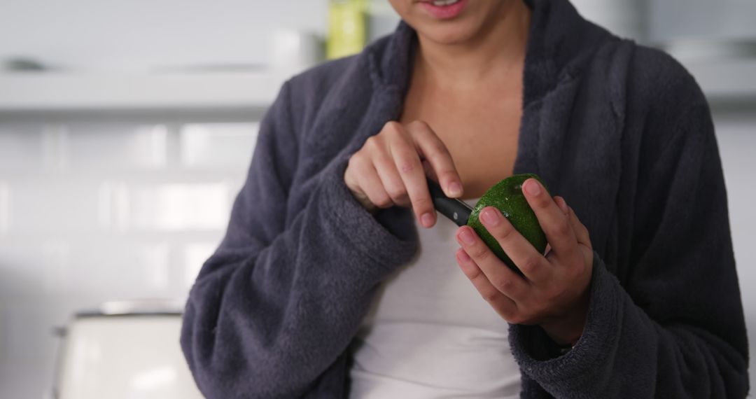 Person Cutting Avocado with Knife Wearing Robe in Modern Kitchen - Free Images, Stock Photos and Pictures on Pikwizard.com