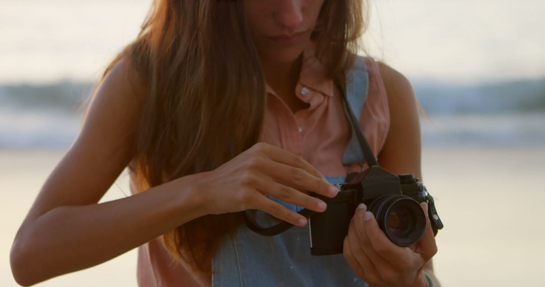 Young Woman Adjusting Camera on Beach at Sunset - Free Images, Stock Photos and Pictures on Pikwizard.com