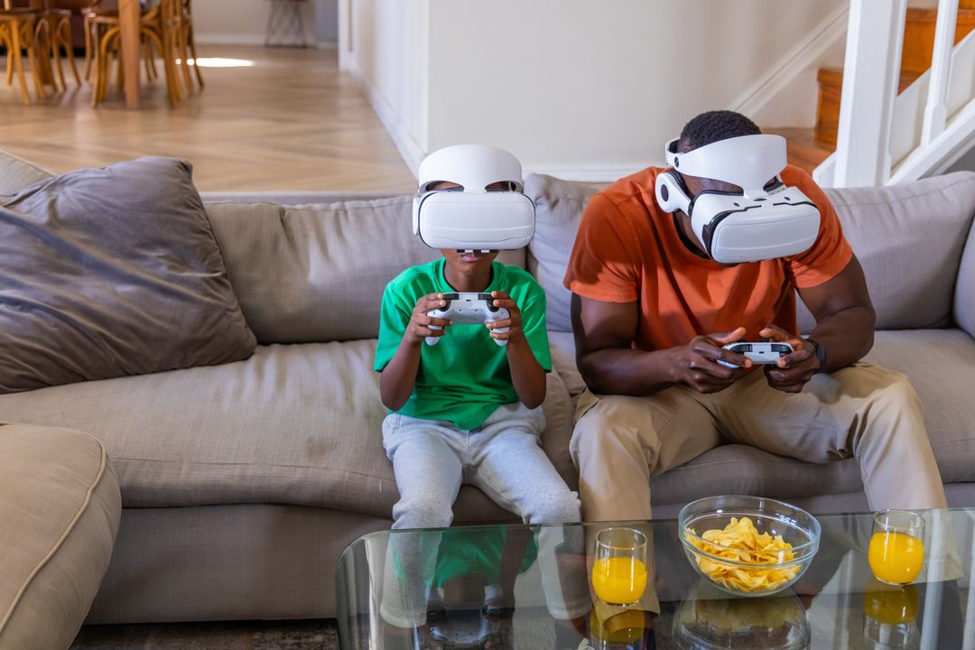 African American Father and Son Playing VR Games on Sofa Wearing Headsets and Using Controllers