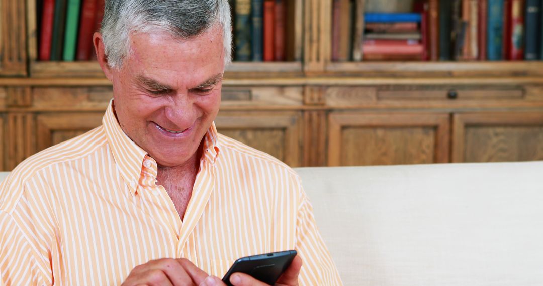 Smiling Senior Man Using Smartphone and Sitting on Couch, Wood Bookshelves - Free Images, Stock Photos and Pictures on Pikwizard.com