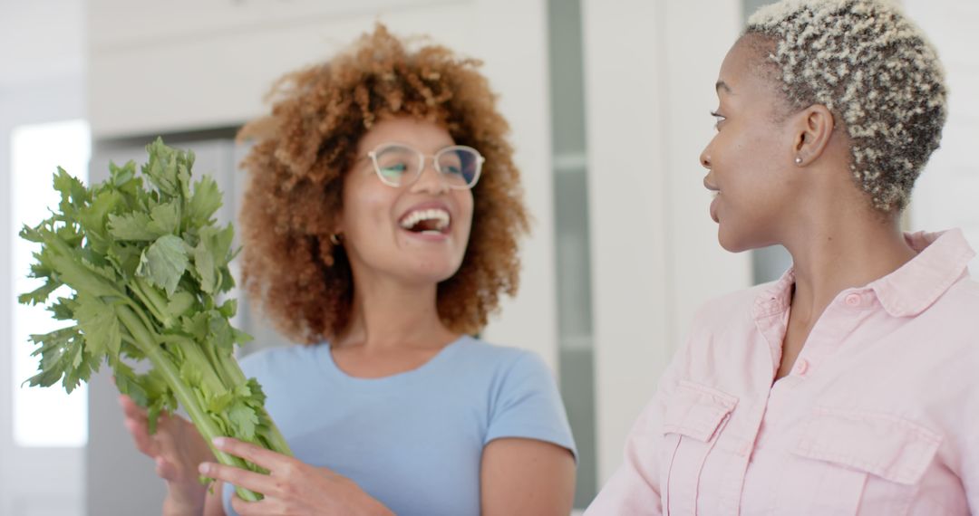 Two Women Laughing Together Holding Fresh Celery in Modern Kitchen - Free Images, Stock Photos and Pictures on Pikwizard.com