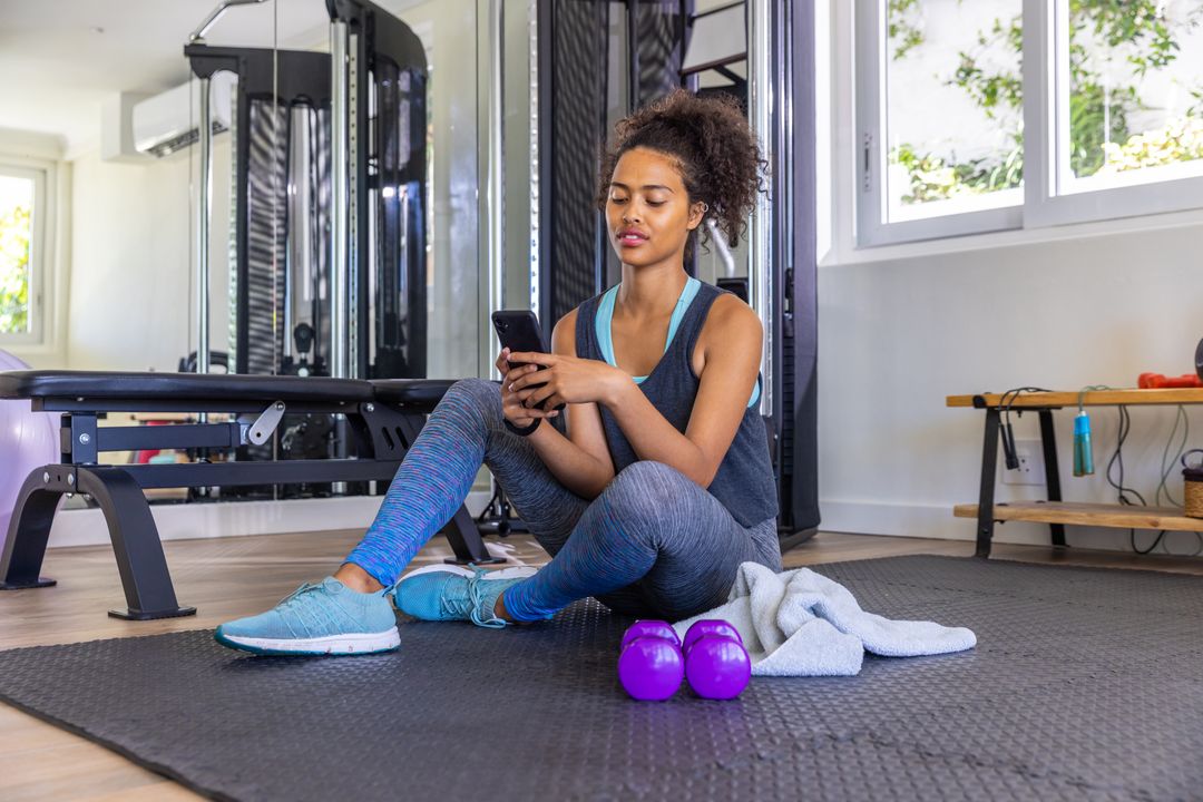 Young Woman Relaxing in Gym with Smartphone and Weights