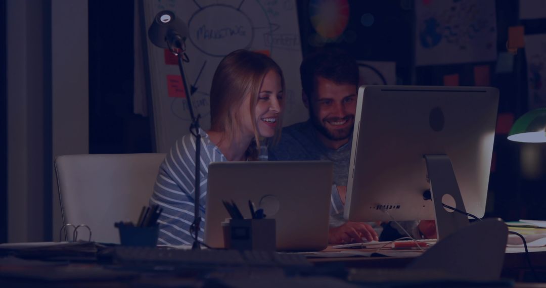 Late night startup collaboration at desk with laptop and desktop monitor, smiling team