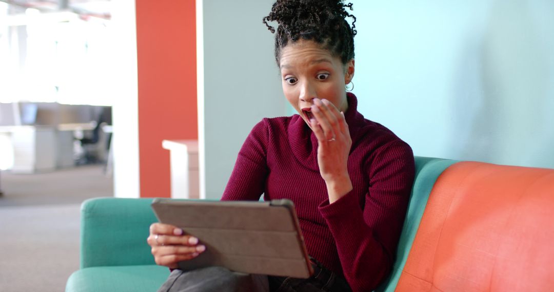 Surprised woman reacting to tablet while sitting on modern two-tone sofa in coworking