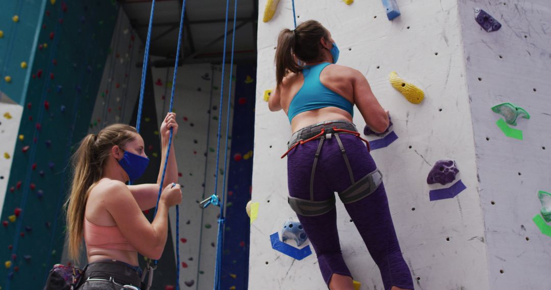Two caucasian women wearing face masks using ropes to climb wall at indoor climbing wall - Free Images, Stock Photos and Pictures on Pikwizard.com