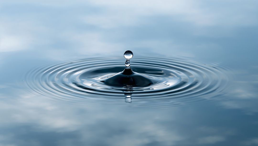 Rising water droplet forming glass bead and concentric ripples on calm blue surface