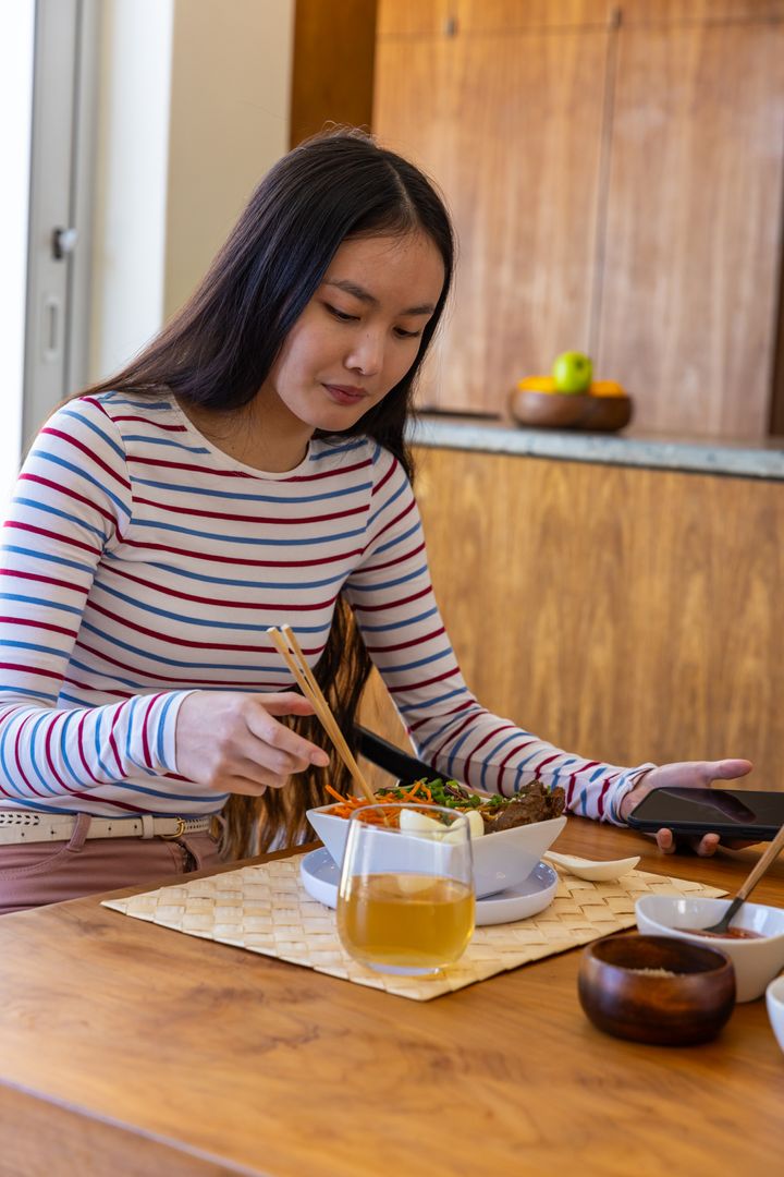Young Woman Enjoying Noodle Dish While Using Smartphone