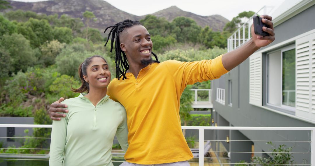 Diverse couple taking selfie on modern balcony surrounded by greenery and mountain views
