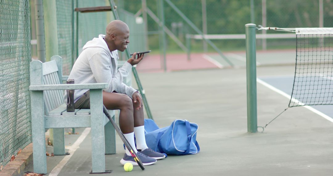 Happy african american male tennis player sitting on bench and talking on smartphone on sunny court - Free Images, Stock Photos and Pictures on Pikwizard.com