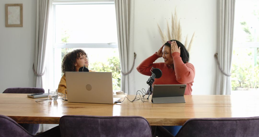 Female colleagues recording podcast at home dining table with laptop and tablet