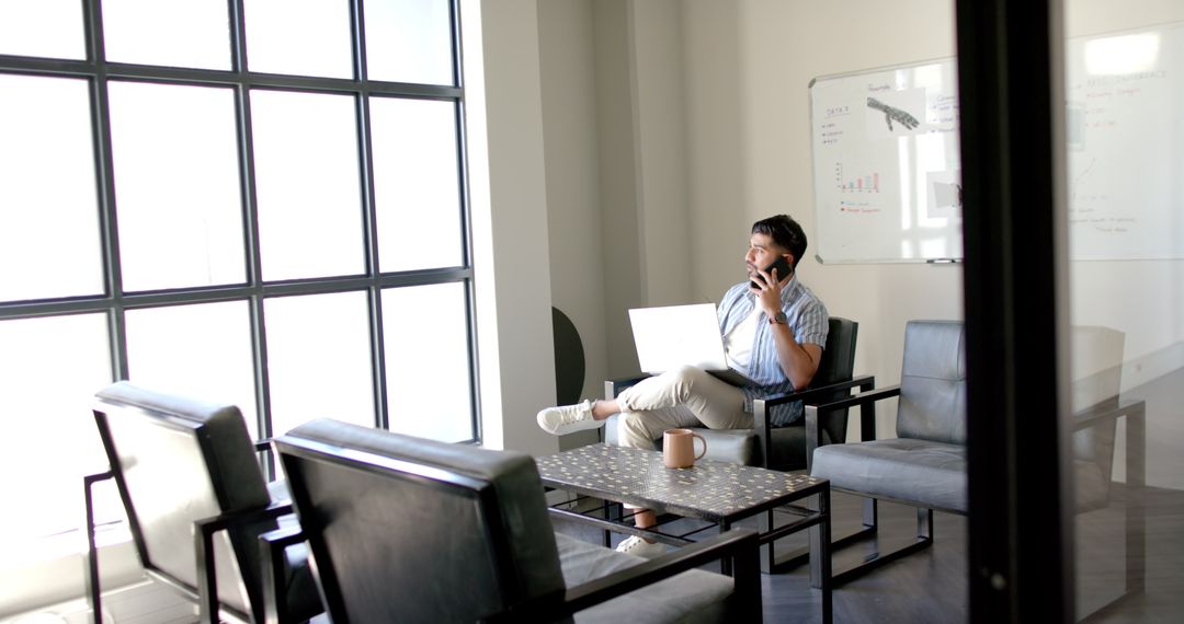 Man Engaged in Phone Call and Laptop Work in Bright Office