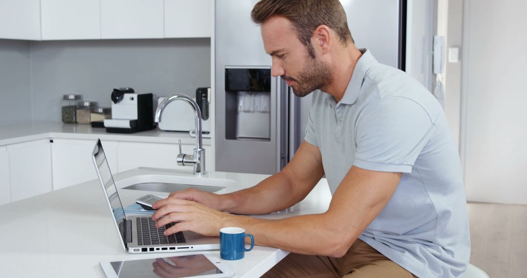 Man Working on Laptop in Modern Kitchen with Tablet and Coffee - Free Images, Stock Photos and Pictures on Pikwizard.com