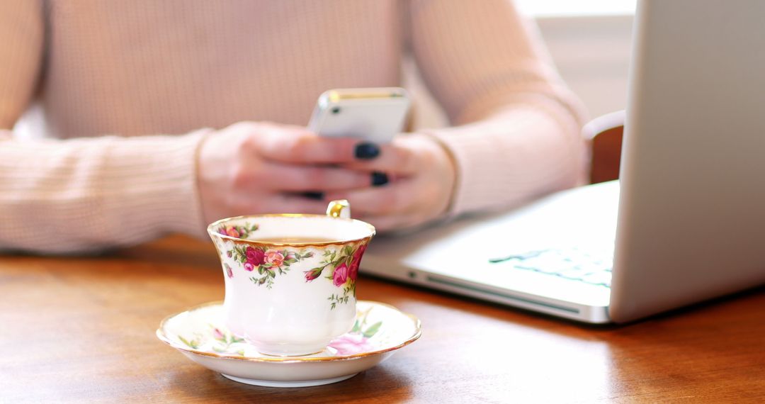 Person Texting on Smartphone Near Laptop with Floral Teacup - Free Images, Stock Photos and Pictures on Pikwizard.com