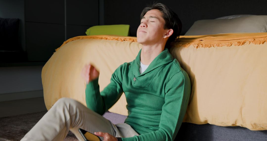 Relaxed Asian Man Lounging Against Bed Using Smartphone