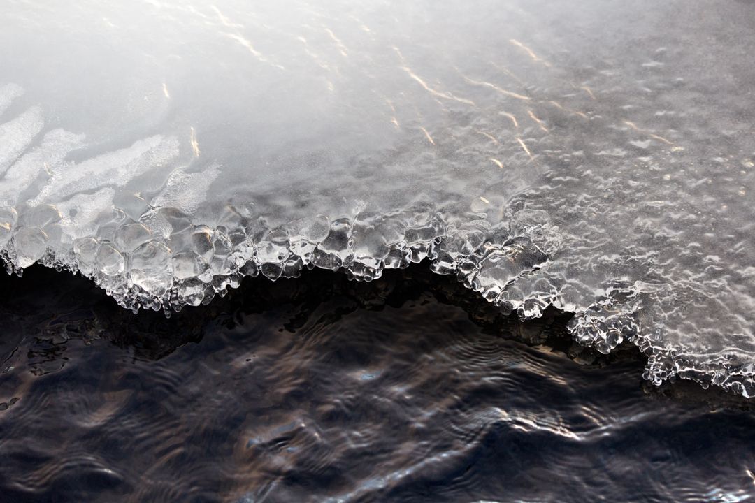 Crystal ice edge melting into dark flowing river water with ripples and reflections