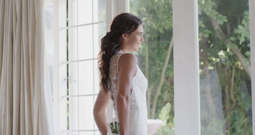 Bride Smiling by Window with Bouquet in Elegant Bridal Dress