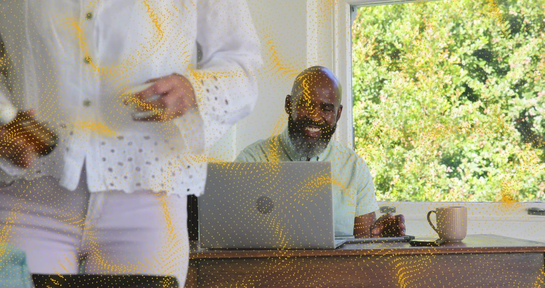 Smiling man working on laptop at home office while senior woman walking past sunny window