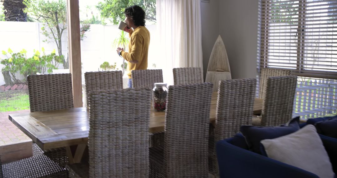 Man Standing by Sliding Door Drinking Coffee While Holding Phone in Sunlit Dining Room