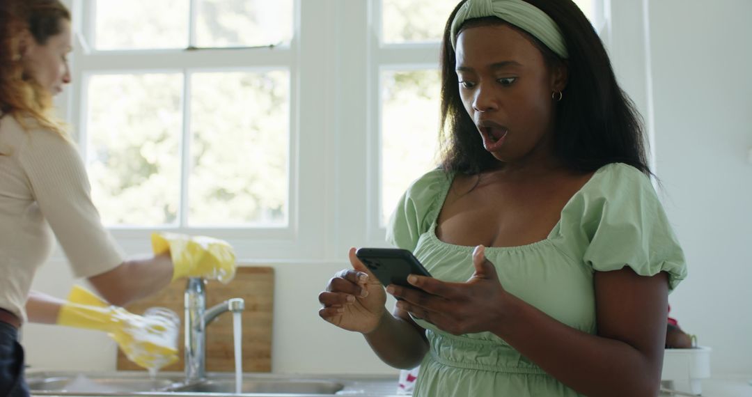 Surprised woman reading smartphone while friend washes dishes
