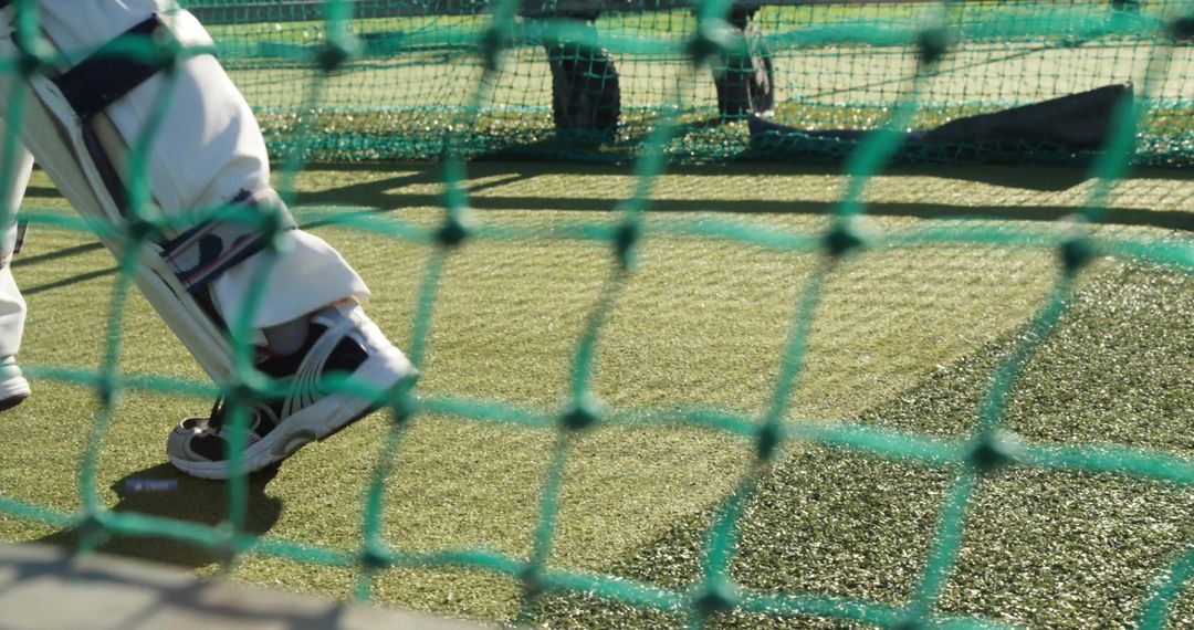 Batsman practicing footwork in net wearing batting pads on sunlit artificial turf