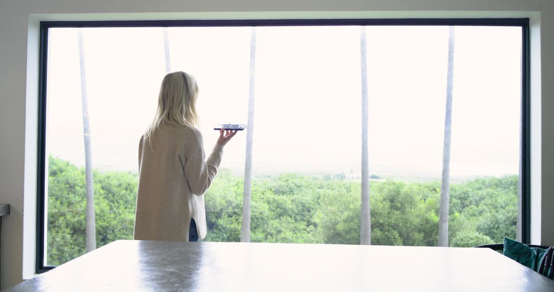 Woman holding smartphone at large window overlooking green palms and coastal panorama