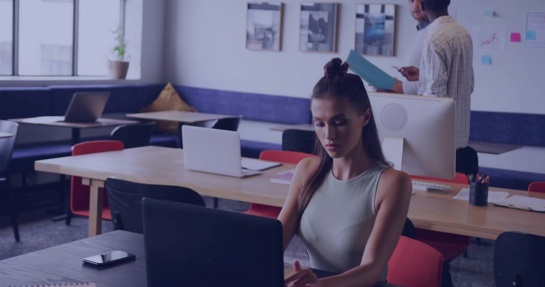 Focused woman typing on laptop in modern coworking space with smartphone and team collaboration