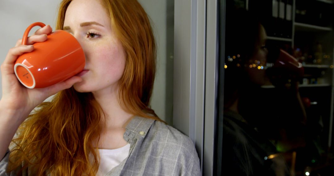 Red-haired woman drinking coffee by window at night - Free Images, Stock Photos and Pictures on Pikwizard.com