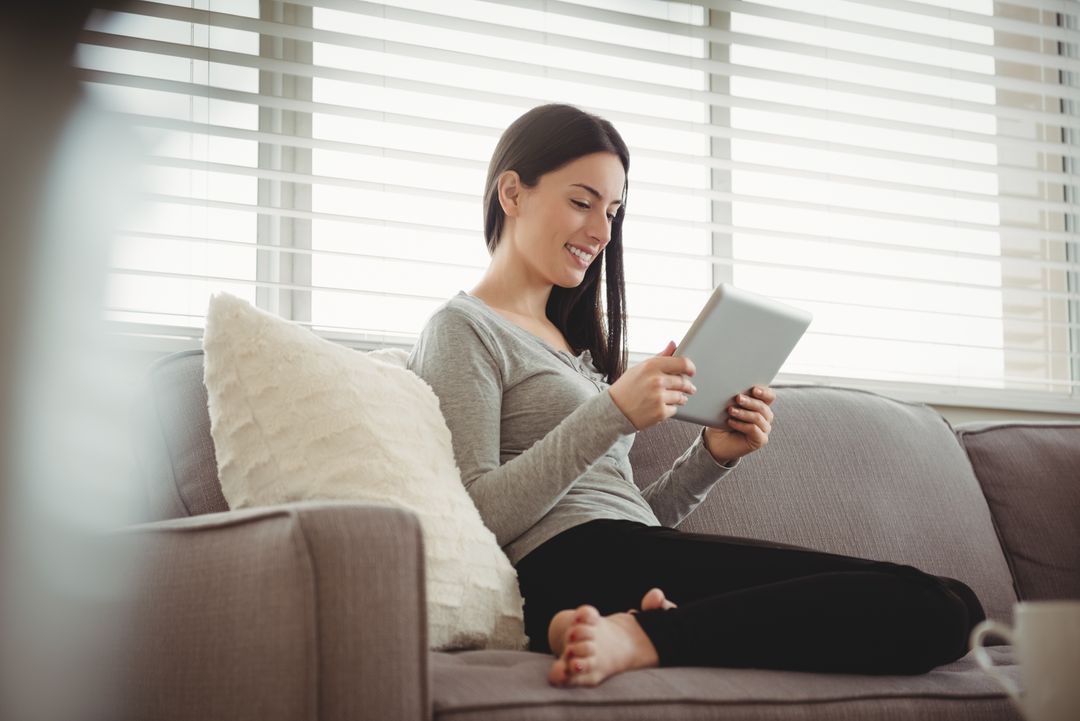 Smiling woman holding tablet while sitting on sofa - Free Images, Stock Photos and Pictures on Pikwizard.com