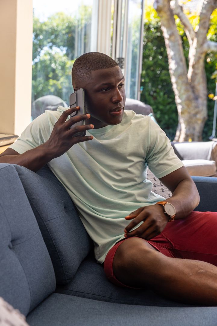 African American Man Talking on Smartphone While Relaxing on Sofa by Glass Door in Casual Outfit