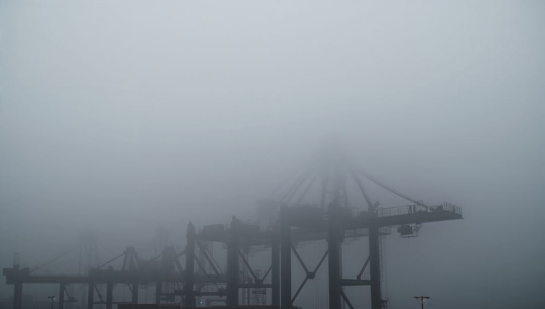 Gantry cranes emerging from dense fog at harbor terminal, moody industrial skyline silhouettes