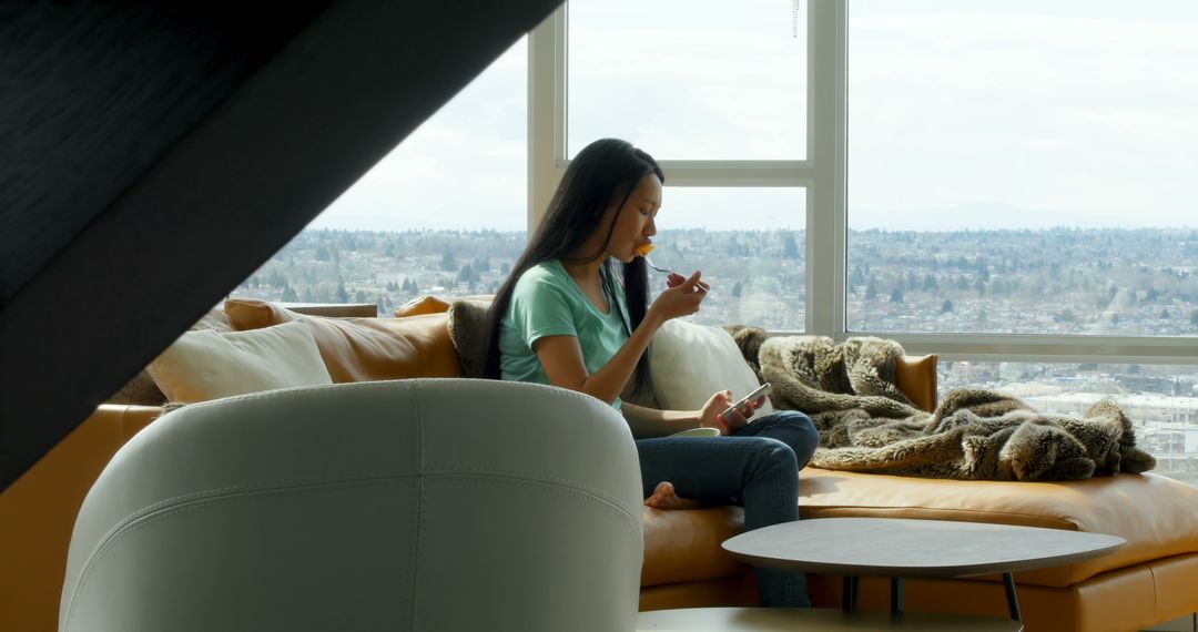 Woman Relaxing on Sofa by Window Eating Snack in Modern Urban Apartment - Free Images, Stock Photos and Pictures on Pikwizard.com