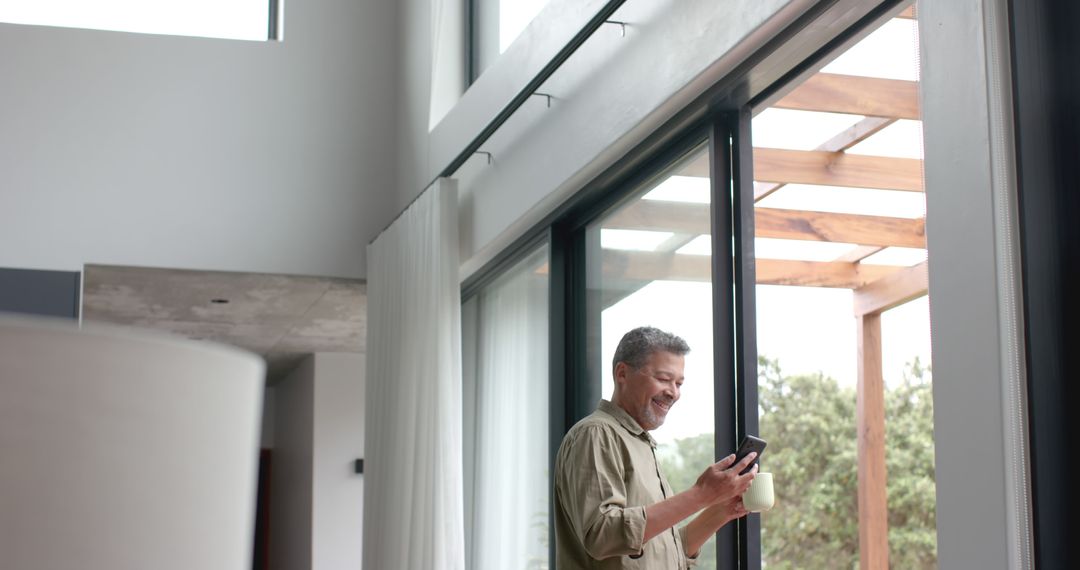 Middle-aged man enjoying coffee while using smartphone near large window - Free Images, Stock Photos and Pictures on Pikwizard.com