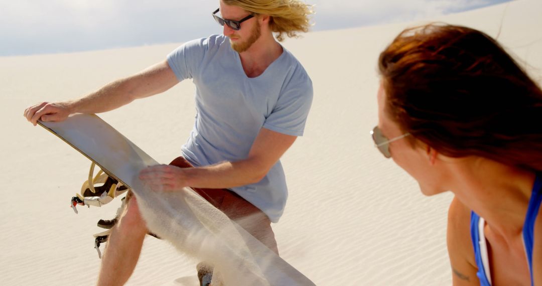 Man Preparing Sandboard at Desert Location with Woman Watching