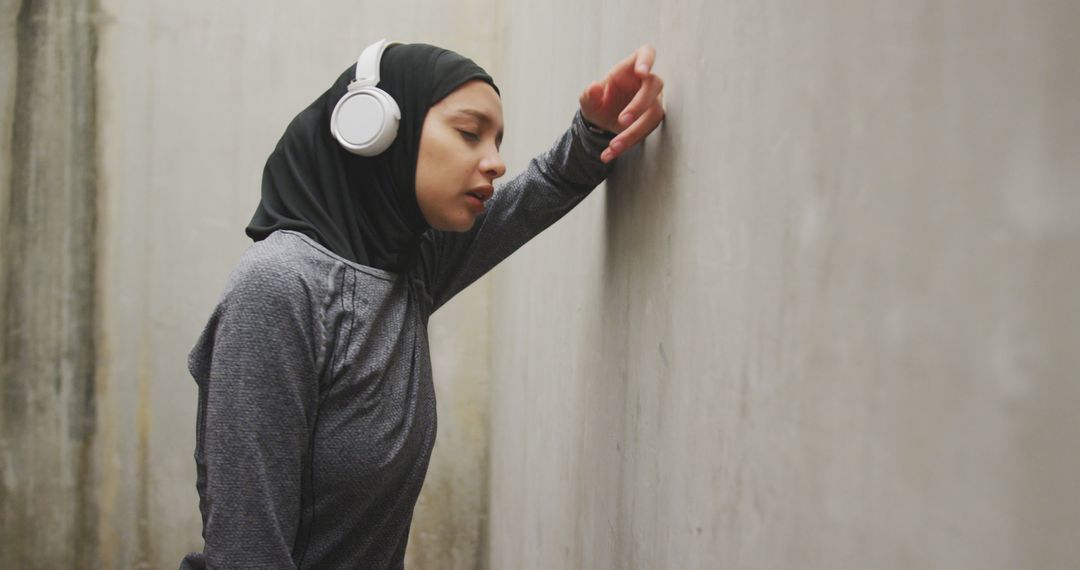 Tired Athlete in Hijab with Headphones Resting Against Wall