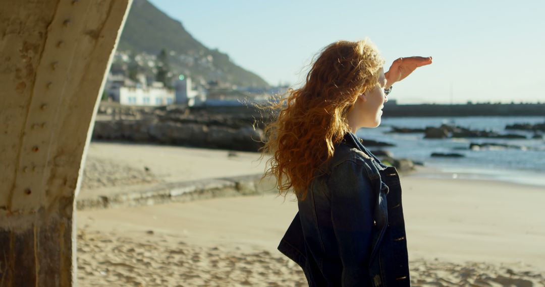 Redhead woman shielding eyes while looking at sea on sunny beach - Free Images, Stock Photos and Pictures on Pikwizard.com