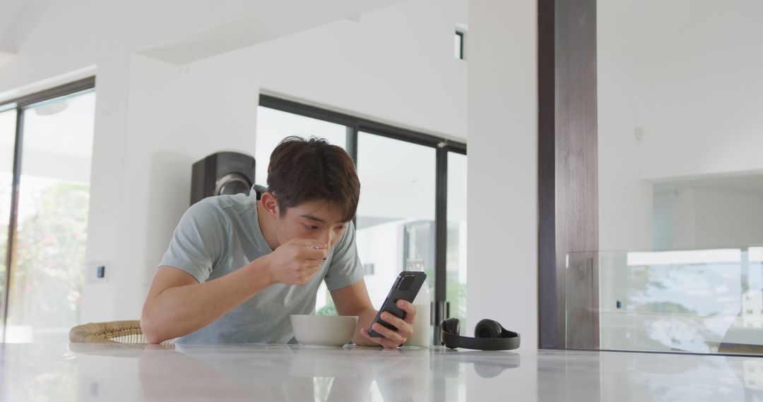 Young Man Eating Breakfast While Using Smartphone in Modern Kitchen - Free Images, Stock Photos and Pictures on Pikwizard.com